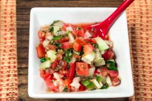 Small square white bowl Garden Salsa Fresca is a naturally sos free salsa fresca with fresh tomatoes, white onion, cucumbers, cilantro, burst of lime juice and a little kick from the jalapeno pepper. with a red spoon.