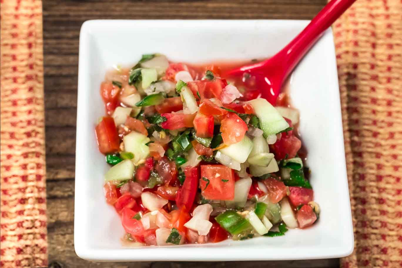 Small square white bowl Garden Salsa Fresca is a naturally sos free salsa fresca with fresh tomatoes, white onion, cucumbers, cilantro, burst of lime juice and a little kick from the jalapeno pepper. with a red spoon.