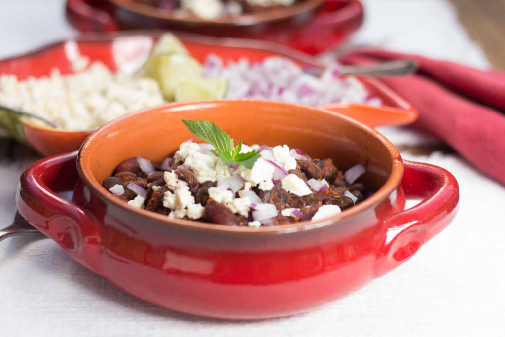 Red-glazed terracotta casserole serving dish with Chile Bean ChiliOil-free Vegan Chile Bean Chili and a chile pepper-shaped dish with tangy bites, lime wedges and diced red onion behind.