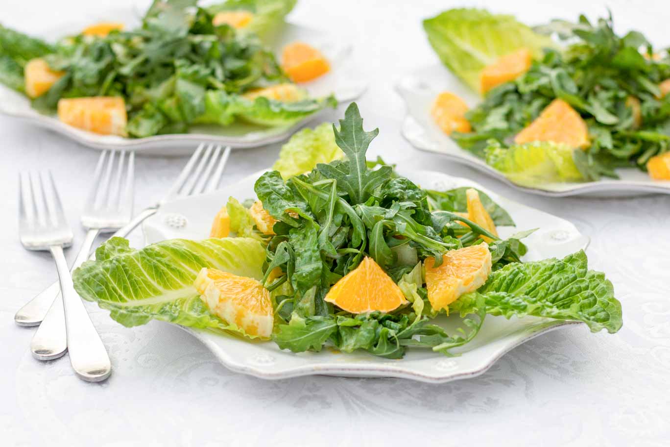 Three white scalloped plates with Whole Orange Balsamic Arugula Salad on a white tablecloth.