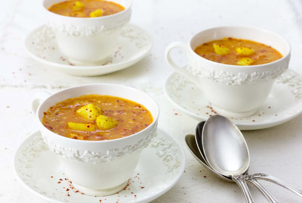 Three white wedgewood cups of Red Lentil Soup with Carrot and Ginger garnished with spice potatoes and a sprinkle of Aleppo pepper.