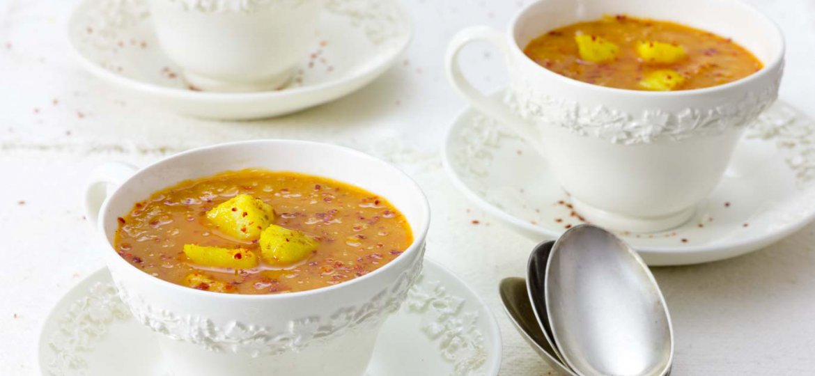 Three white wedgewood cups of Red Lentil Soup with Carrot and Ginger garnished with spice potatoes and a sprinkle of Aleppo pepper.