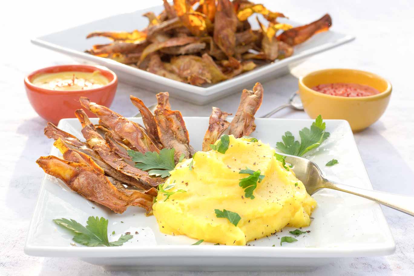 White square plate with coil-free potato skin crisps and golden mashed potatoes. Two condiment dishes behind and a platter of the oil-free potato skin crisps.