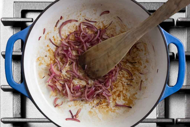 Sliced onions browning on medium heat in a cast iron enameled braiser pan with a wood spatul