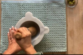 Crushing garlic with a wood pestle in a marble mortar placed on a cutting board with a towel under.