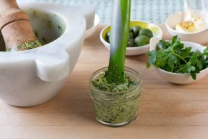 Oil-free vegan parsley pesto in a small glass jar with flexible spatula. Marble mortar and wood pestle to the left and small bowls of olives, garlic and parsley behind.