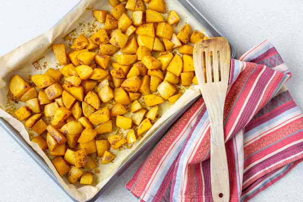 turmeric spiced oil-free roasted potatoes on a baking sheet with a tea towel and wooden spatula on the side