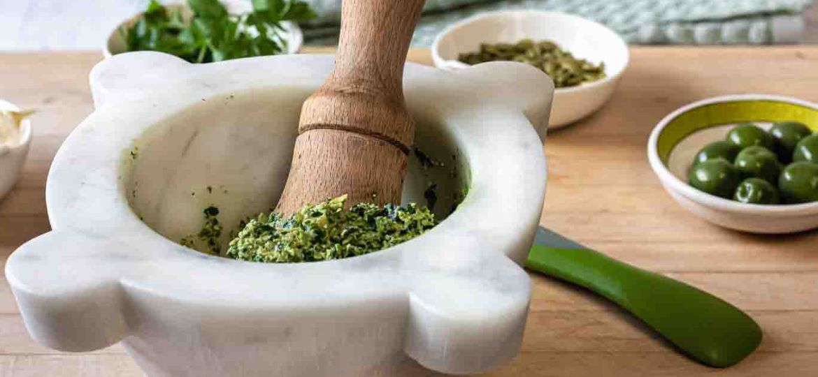 Oil-free Vegan Parsley pesto in the marble mortar with the wood pestle on a cutting board small bowls of Castelvetrano olives, pumpkin seeds and parsley leaves.